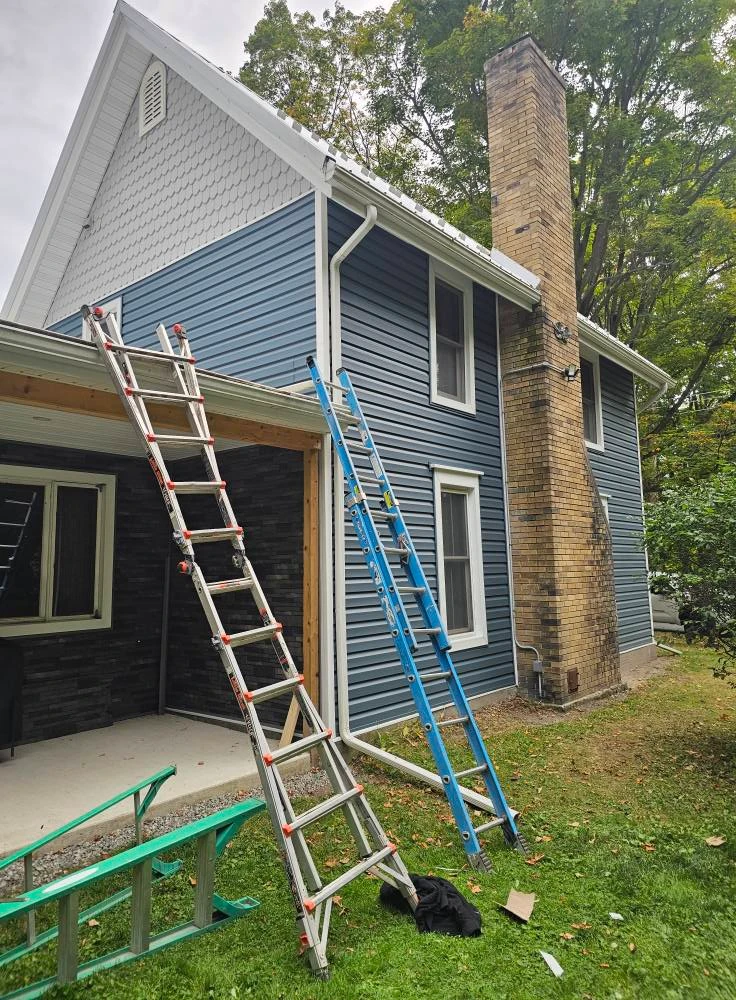 Two story home mid re-side with new blue vinyl siding, brick chimney and ladders staged against the wall