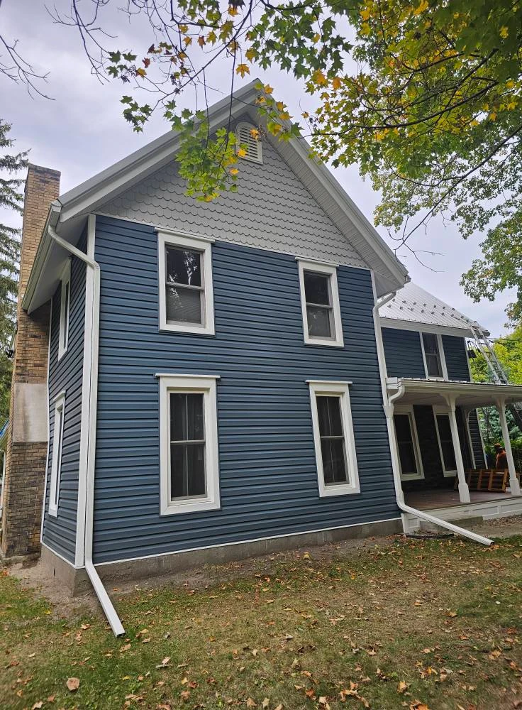 Finished side elevation of a farmhouse with blue vinyl siding, fish scale gable accent, white trim and metal roof