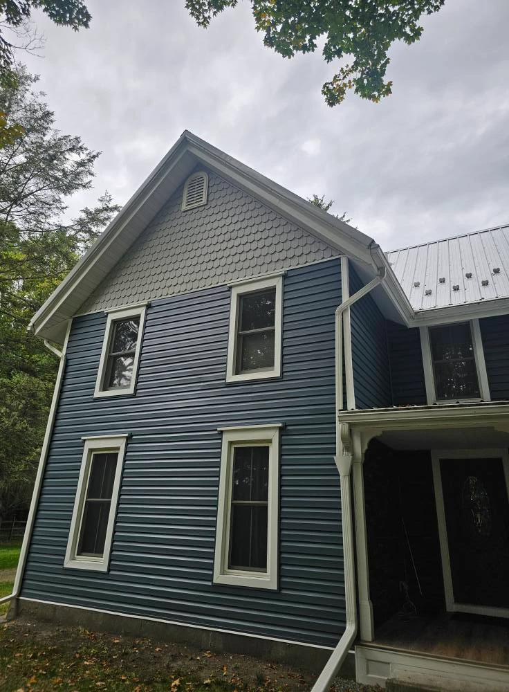 Blue vinyl siding with fish scale shingle gable accent and white window trim on a Finger Lakes farmhouse